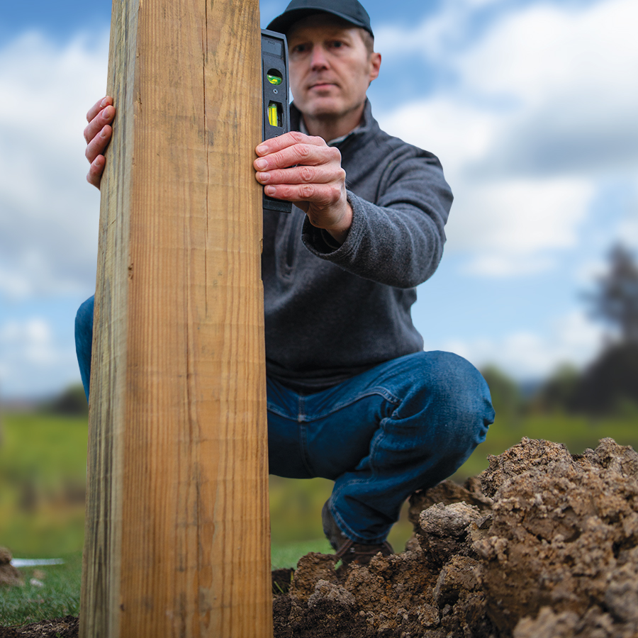 A man using a level to check the alignment of a wood post in the ground.